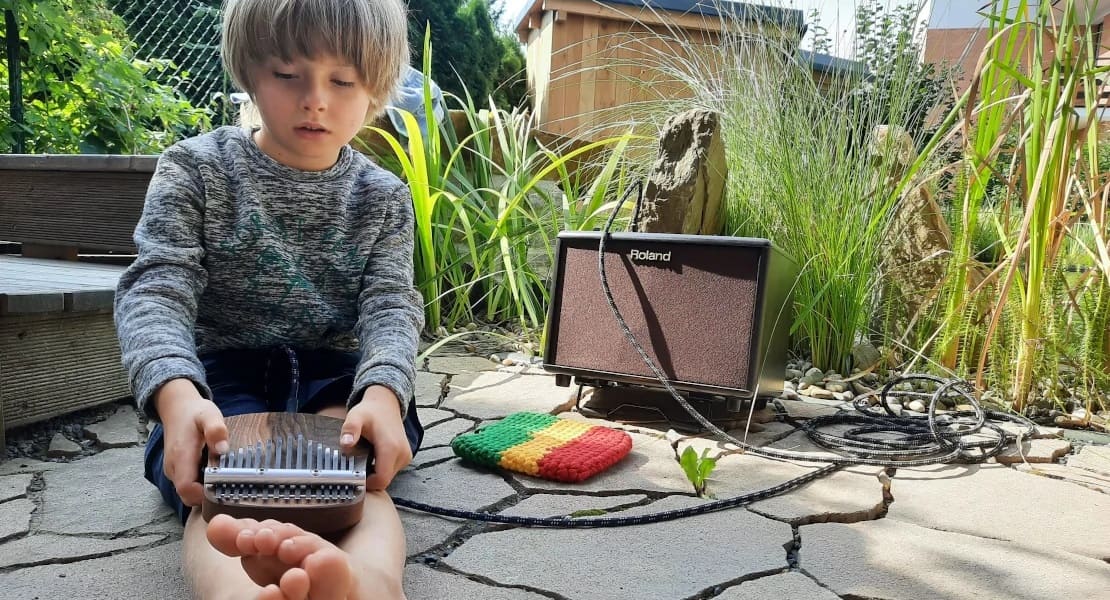A boy sitting on the ground playing a kalimba with a speaker in his yard.