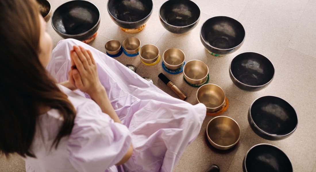 A woman seen from behind meditating surrounded by singing bowls
