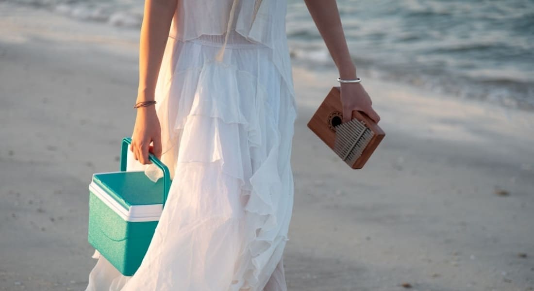 A woman in a white dress carrying a kalimba and a cooler