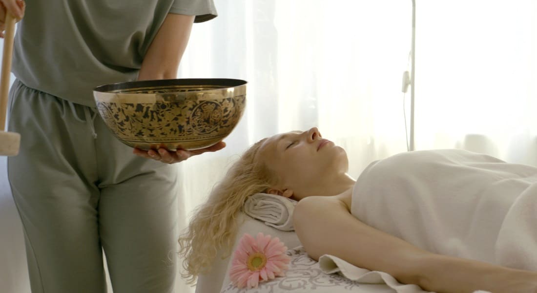 A woman lying down with her eyes closed while another plays a Tibetan singing bowl