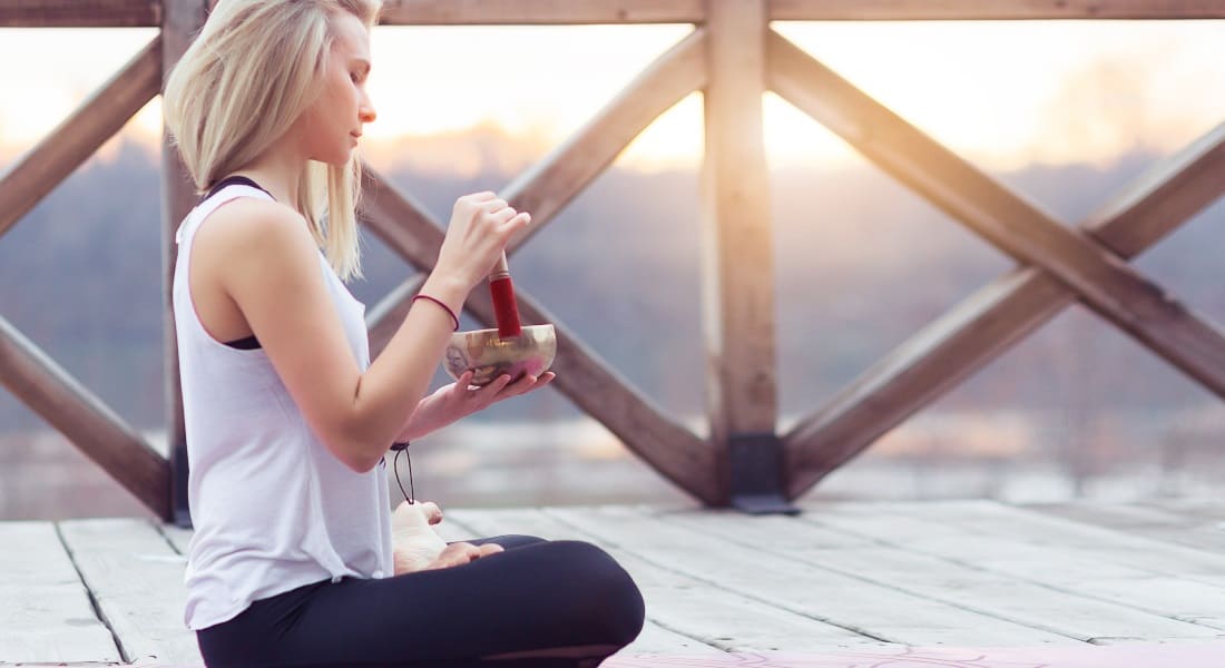 A blonde woman seen from the side sitting in lotus position with a Tibetan singing bowl