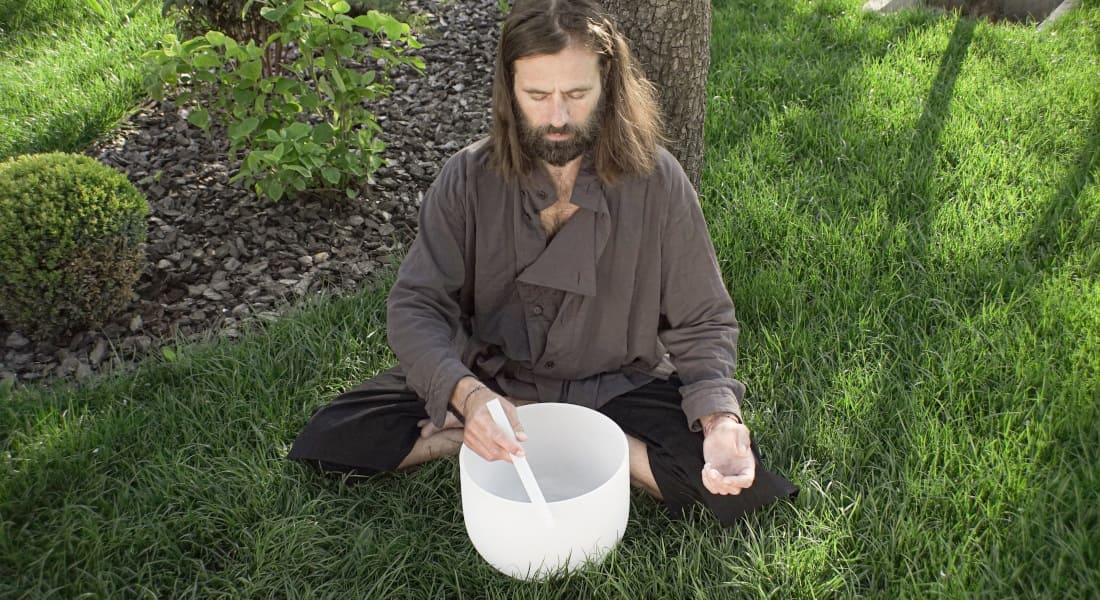 A man sitting in lotus position in a park playing a crystal singing bowl