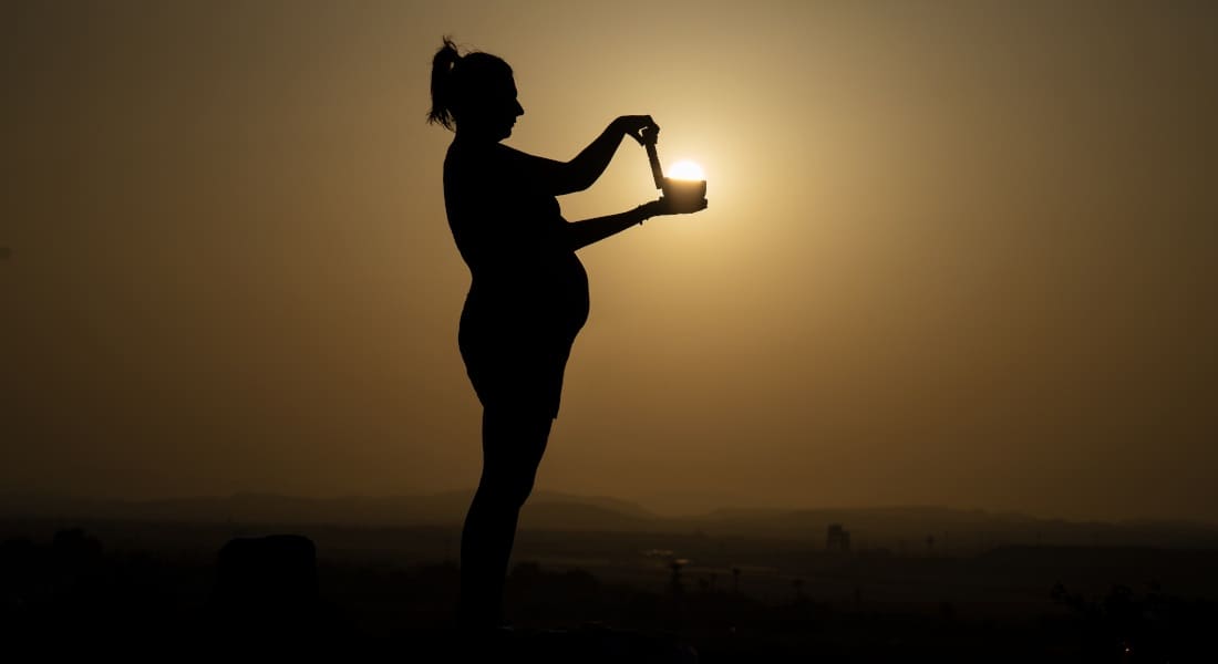 A pregnant woman with a Tibetan singing bowl in front of a sunset