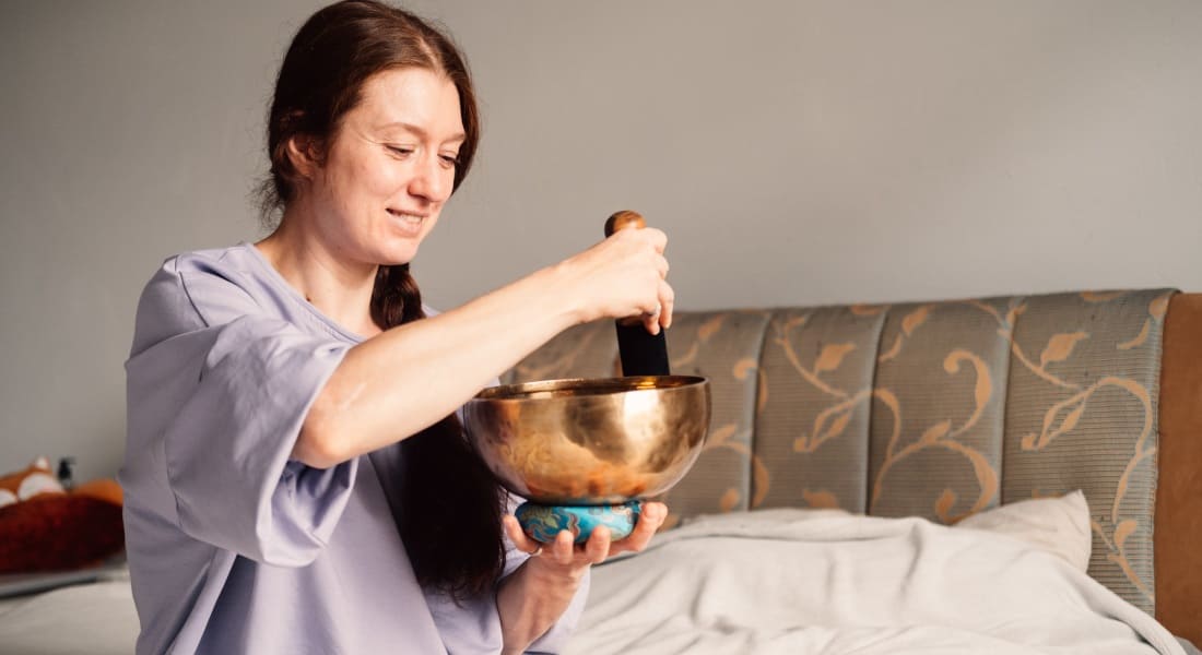 A smiling woman playing a Tibetan singing bowl on her bed