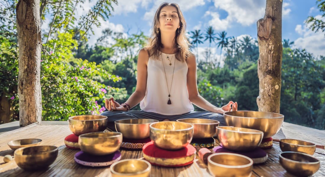 A woman sitting in lotus pose meditating surrounded by singing bowls