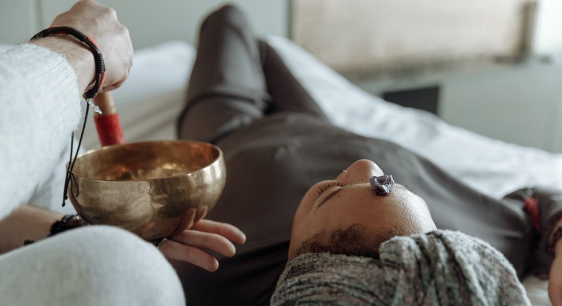 A woman lying down with a stone on her forehead and another person playing a Tibetan singing bowl