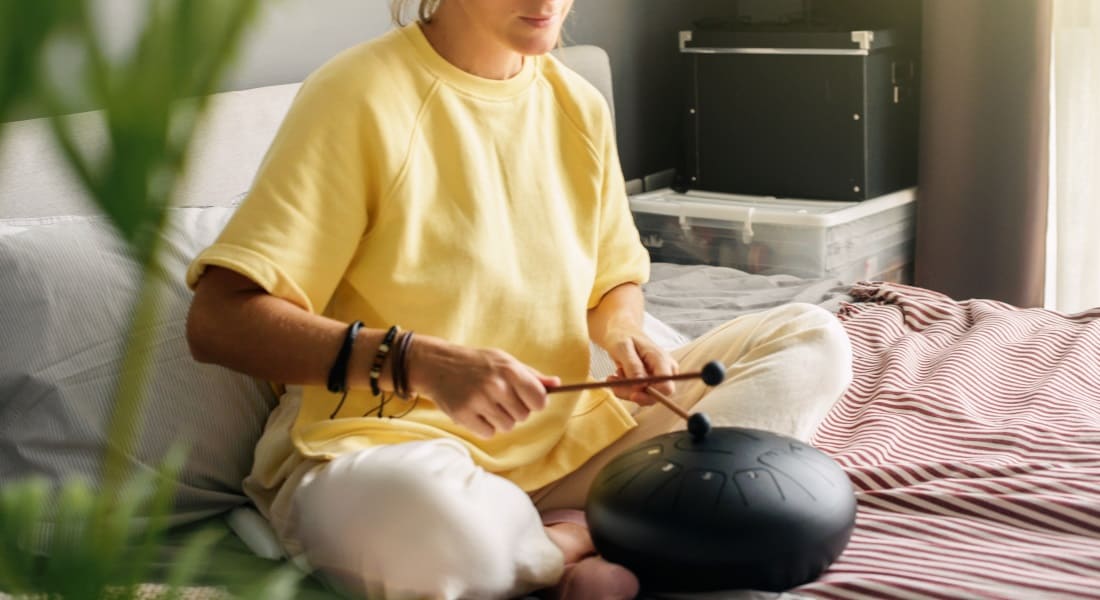 A woman sitting cross-legged on a bed playing a tongue drum