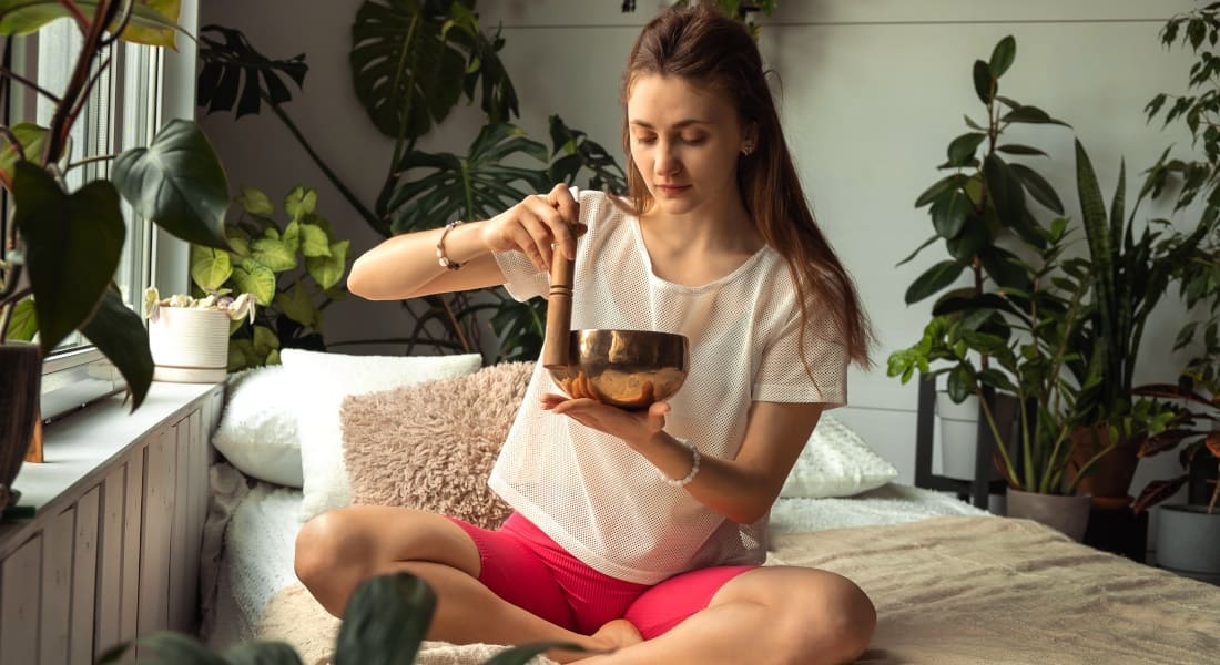 A young woman on a bed surrounded by plants playing a Tibetan singing bowl