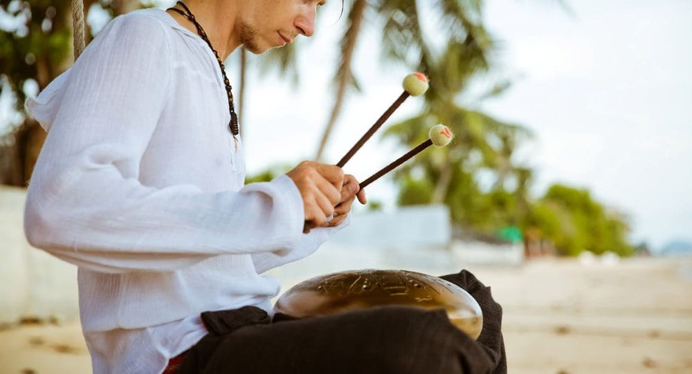 A man sitting on the beach playing a tongue drum with mallets