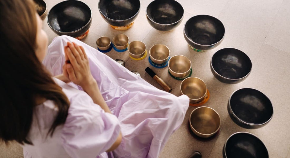 A woman seen from behind meditating surrounded by singing bowls