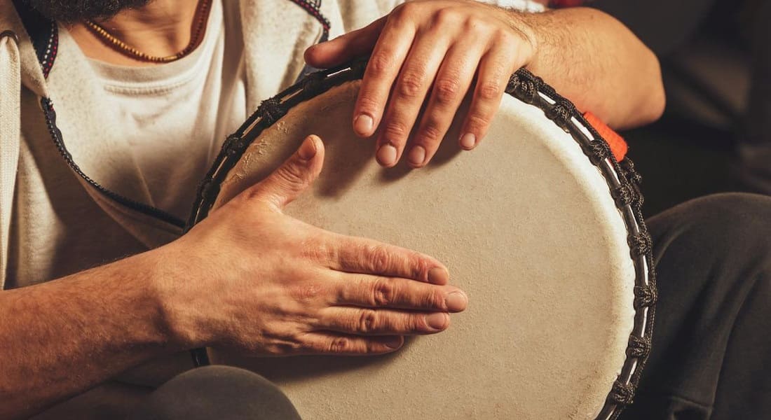 A bearded man playing a djembe while seated