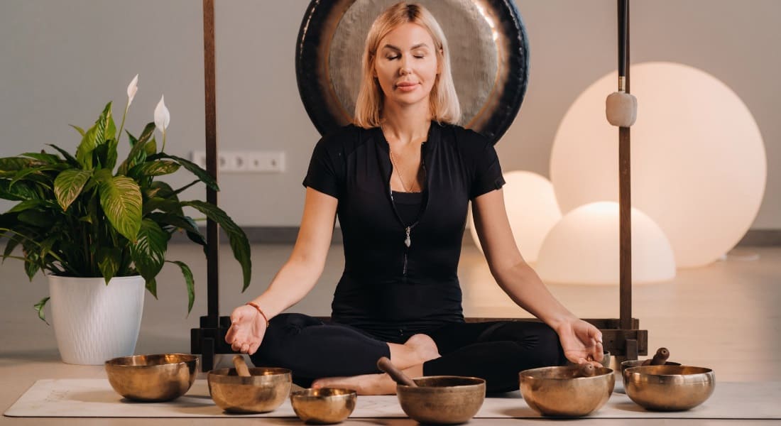 A blonde woman meditating with a gong and Tibetan singing bowls