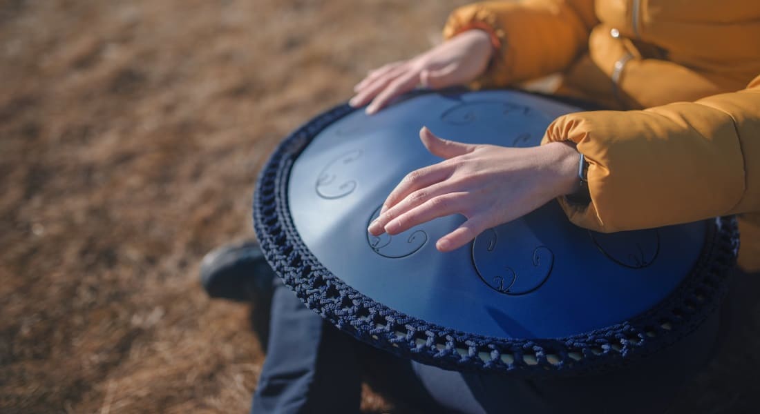 Close-up of a woman's hands playing a blue tongue drum. She is sitting cross-legged in nature.