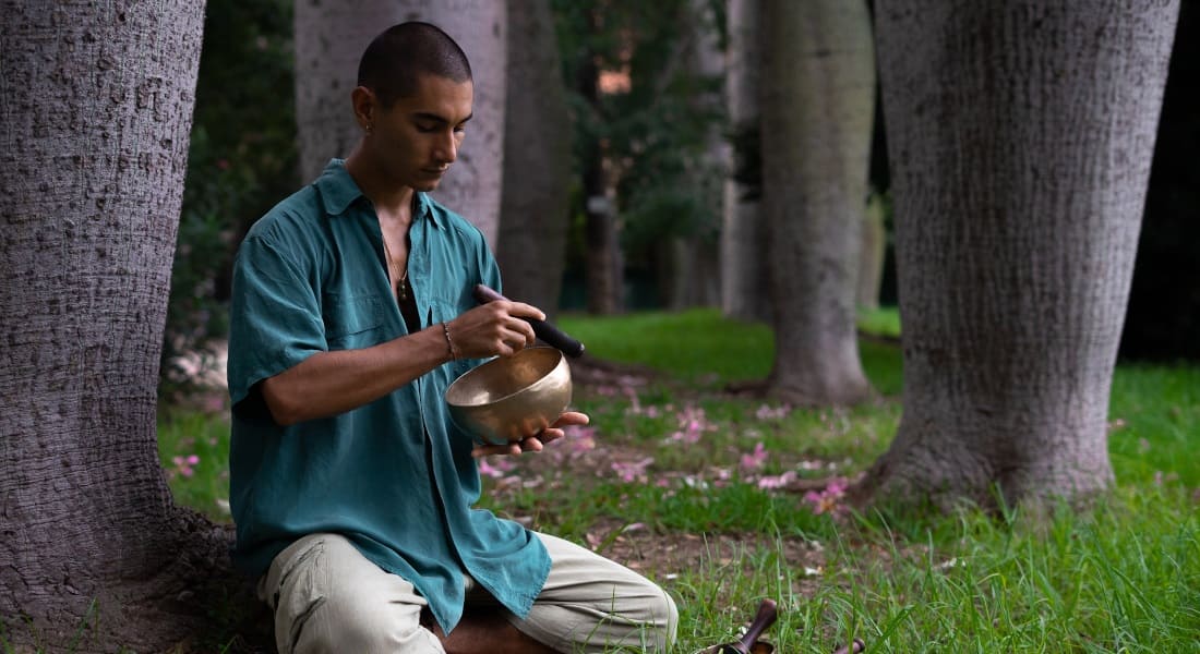 A kneeling man playing a Tibetan singing bowl in the forest
