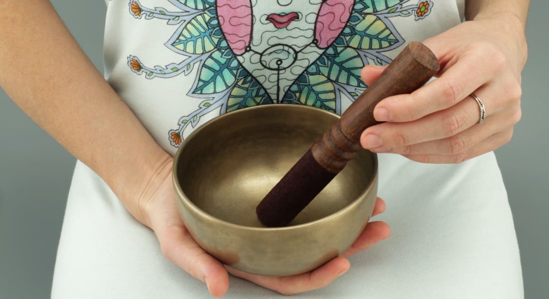 A person holding a metal Tibetan singing bowl in one hand and a mallet in the other