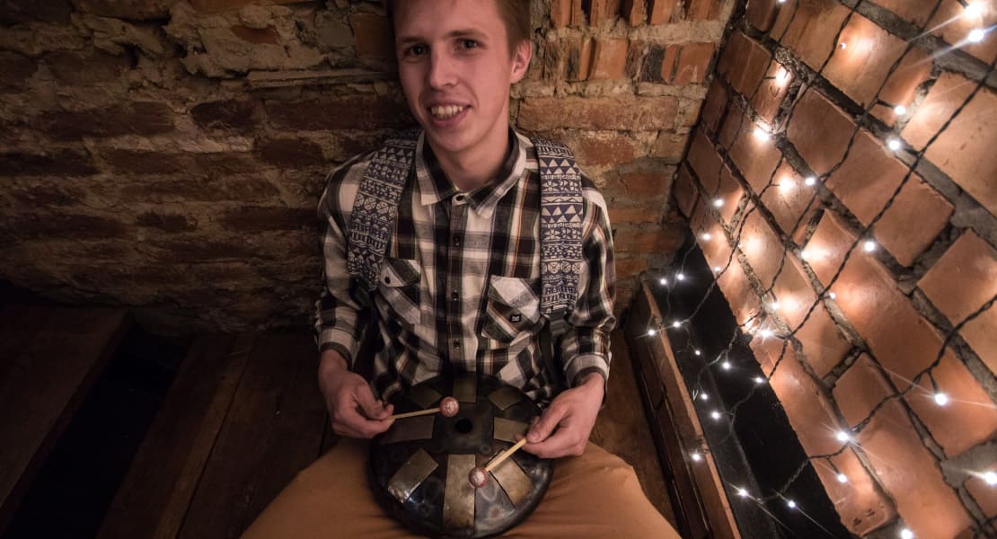 A man sitting on the floor playing a tongue drum and smiling