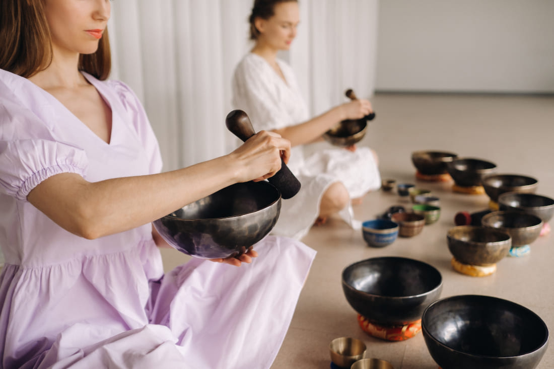 Two women sitting cross-legged playing several Tibetan singing bowls