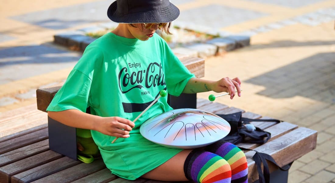 An Asian woman wearing a hat playing a steel tongue drum