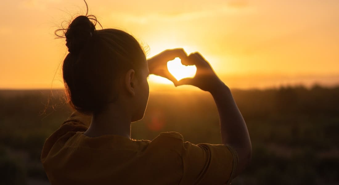 A woman drawing a heart with her hands behind a ray of sunlight