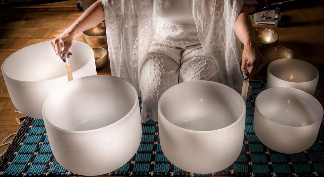 A woman kneeling surrounded by metal and crystal Tibetan singing bowls