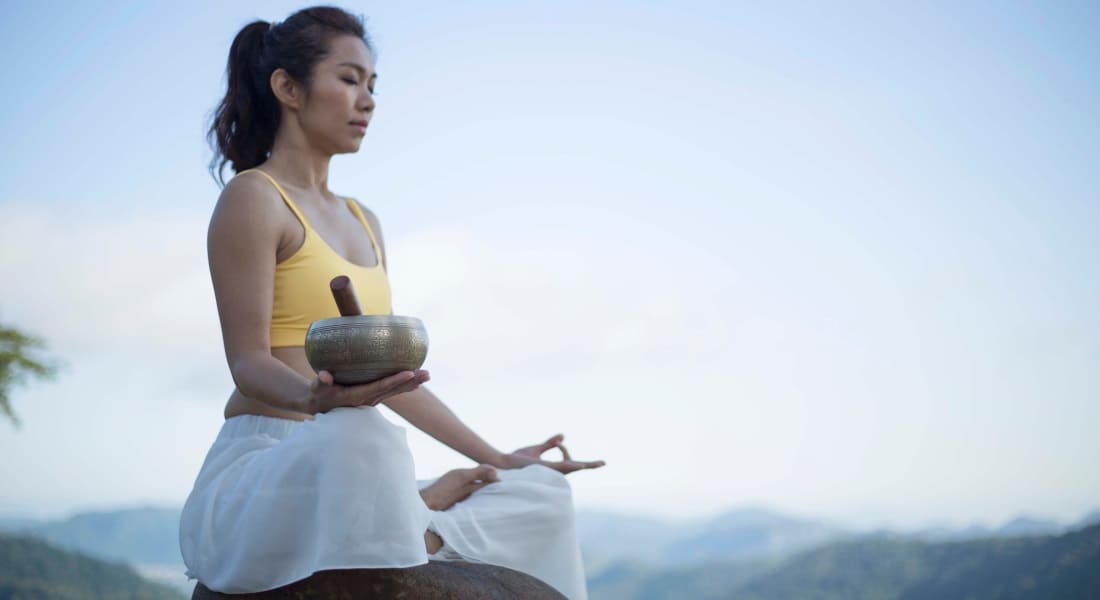 A woman meditating while seated, holding a metal singing bowl in one hand