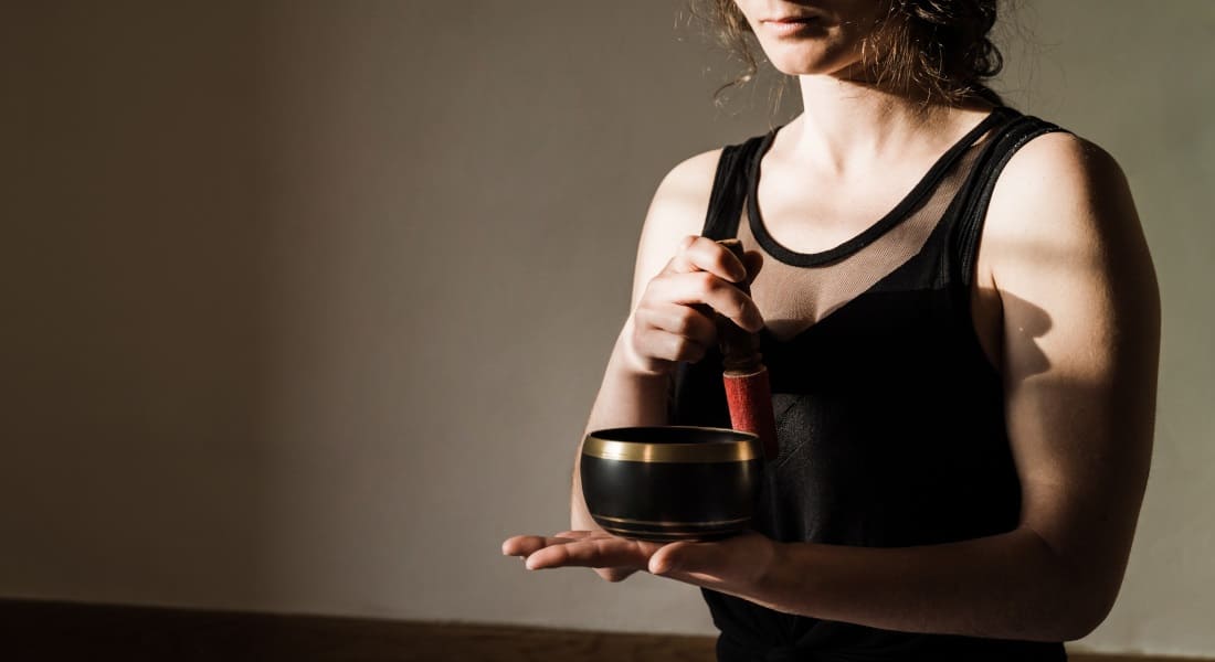 A woman sitting cross-legged holding a black metal Tibetan singing bowl in the palm of her hand.